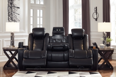 The Party Time power reclining sofa takes its cue from luxury automobiles with lattice- and cross-hatch stitching, for a richly tailored aesthetic that gives you plenty of reasons to celebrate. Sumptuously padded cushions and dramatic midnight black faux leather upholstery add to the indulgence. When it’s time to rev up the action, the sofa’s dual reclining bucket seats (middle seat is stationary), Easy View™ power adjustable headrests and center seat that folds down into a table keep you in the driver’s seat. Dual-sided recliner | One-touch power controls with adjustable positions | Easy View™ power adjustable headrests | Corner-blocked frame with metal reinforced seats | Attached backs and seat cushions | High-resiliency foam cushions wrapped in thick poly fiber | Polyester/polyurethane upholstery | Flip up padded armrests with hidden storage underneath | Drop down table with 2 cup holders and a handy docking station for charging electronics | Includes USB charging port in each power control | Flip up LED light (under center seat headrest) | Ambient blue LED lighting on cup holders and base for a theater-style experience | Power cord included; UL Listed | Estimated Assembly Time: 15 Minutes