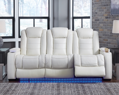 The Party Time power reclining sofa takes its cue from luxury automobiles with lattice and crosshatch stitching, for a richly tailored aesthetic that gives you plenty of reasons to celebrate. Sumptuously padded cushions and crisp white faux leather upholstery add to the indulgence. When it’s time to rev up the action, the dual reclining bucket seats (middle seat is stationary), Easy View™ power adjustable headrests and a center seat that folds down into a table keep you in the driver’s seat. Ambient blue LED lighting on the base and cup holders completes the theater-style experience.Dual-sided recliner | Polyester/polyurethane upholstery | Corner-blocked frame with metal reinforced seat | Attached back and seat cushions | High-resiliency foam cushions wrapped in thick poly fiber | One-touch power control with adjustable positions, Easy View™ adjustable headrest and USB plug-in | Flip up padded armrests with hidden storage | Drop down table with 2 cup holders and a handy docking station for charging electronics | Flip up LED light (under center seat headrest) | LED lighting on cup holders and base for theater-style experience | Power cord included; UL Listed | Estimated Assembly Time: 15 Minutes