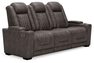 The Hyllmont power reclining sofa in trendy gray is proof of just how far recliners have come when it comes to form and function. Ultra cool and contemporary, this fully loaded power reclining sofa makes it so easy to social distance and make home your haven. Dressed to impress with diamond stitching inspired by sports car interiors, this reclining sofa is covered in a fabulous faux leather with pebbled effect for authenticity. High-tech advancements include a one-touch power control with Easy View™ adjustable headrest and USB plug-in and a drop-down table with cup holders, flip-up light and AC power/USB plug-ins. Dual-sided recliner | One-touch power control with adjustable positions, Easy View™ adjustable headrest and USB plug-in | Corner-blocked frame with metal reinforced seat | Attached cushions | High-resiliency foam cushions wrapped in thick poly fiber | Each armrest with hidden storage and cup holder | Drop-down center table with 2 cup holders, flip-up light and hidden AC power and USB ports | Faux leather polyester upholstery | Power cord included; UL Listed | Exposed tapered feet | Estimated Assembly Time: 15 Minutes