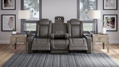 The Hyllmont power reclining sofa in trendy gray is proof of just how far recliners have come when it comes to form and function. Ultra cool and contemporary, this fully loaded power reclining sofa makes it so easy to social distance and make home your haven. Dressed to impress with diamond stitching inspired by sports car interiors, this reclining sofa is covered in a fabulous faux leather with pebbled effect for authenticity. High-tech advancements include a one-touch power control with Easy View™ adjustable headrest and USB plug-in and a drop-down table with cup holders, flip-up light and AC power/USB plug-ins. Dual-sided recliner | One-touch power control with adjustable positions, Easy View™ adjustable headrest and USB plug-in | Corner-blocked frame with metal reinforced seat | Attached cushions | High-resiliency foam cushions wrapped in thick poly fiber | Each armrest with hidden storage and cup holder | Drop-down center table with 2 cup holders, flip-up light and hidden AC power and USB ports | Faux leather polyester upholstery | Power cord included; UL Listed | Exposed tapered feet | Estimated Assembly Time: 15 Minutes