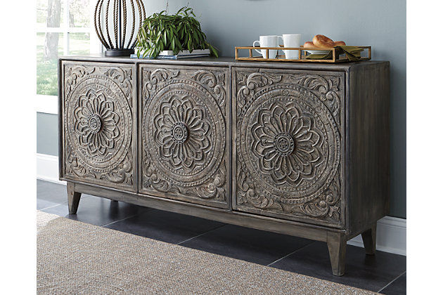 Have a taste for something exotic? Feast your eyes on the exquisitely styled Fair Ridge console. Whether tantalizing in the dining room, gracing an entryway or serving as an eclectic TV stand—what a masterpiece of an accent piece. Fancifully floral-carved doors are a striking contrast to the console’s ultra-linear profile.Made of solid wood | Hand-carved accents | 3 shelves behind 3 cabinet doors | Antique gray finish | Includes tipping restraint pack | Excluded from promotional discounts and coupons | Estimated Assembly Time: 15 Minutes