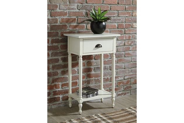 The perfect complement to any sofa, the Juinville accent table embodies quintessential vintage charm. It’s casually cool with a bronze-tone cupped drawer pull, turned legs, a framed drawer front and a classic white finish. You’ll love how the convenient drawer stores remotes and catchalls with ease. Last, but certainly not least, the bottom shelf is a hot spot for decor accents or an extra bin for tip-top organization in small spaces.Made of veneers, wood and engineered wood | White finish | 1 smooth-gliding drawer, 1 lower shelf | Antique bronze-tone finished metal drawer pull | Assembly required | Estimated Assembly Time: 15 Minutes