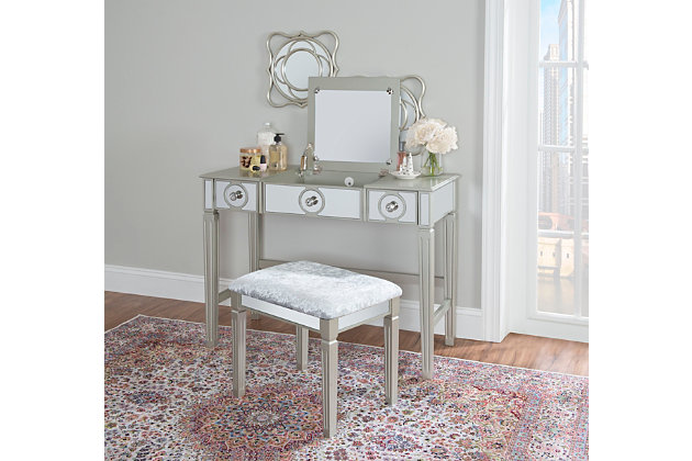 Add a glamourous touch to the room or dressing area with the Renay vanity set. Finished in silver and designed in grand elegance, you'll discover attractive elements like tapered legs with reeds and a mirrored front accented by round silver pulls. The matching stool also has mirrored details and its padded seat is upholstered in gray fabric upholstery. The vanity's mirror can be flipped down when not in use providing you with tabletop space. Plus, three drawers offer hidden storage—perfect for keeping the room tidy.Made of rubberwood, engineered wood and mirror | Silver finish with mirrored front | Flip top mirror | 3 drawers with round silvertone pulls | Includes padded stool upholstered in gray fabric | Tapered, reeded legs | Assembly required