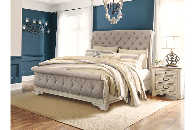 Elevating the art of traditional cottage styling, the Realyn California king upholstered sleigh bed is your dream bedroom retreat realized. A curvaceous beauty, this brilliant upholstered bed wows with a distressed chipped white finish for heirloom appeal. Deep tufting on the cushioned upholstered headboard and footboard adds such a cozy element sure to put you at ease. Mattress and foundation/box spring available, sold separately.Made of wood and engineered wood | Includes headboard, footboard and rails | Distressed, chipped white finish | Polyester upholstery with foam cushion and button tufting | Assembly required | Foundation/box spring required, sold separately | Mattress available, sold separately | Estimated Assembly Time: 70 Minutes