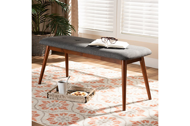 Featuring a stunning medium oak finish, the Flora dining bench brings warmth to any dining space. Constructed from solid rubberwood, it's upholstered in a soft, durable polyester fabric and padded with foam. Expert craftsmanship is displayed in the curved edges and the smooth grained finish. Designed for everyday use, the seat is supported by four angled, tapered legs that create a modern profile while also providing ample legroom. A single row of button tufting adds a decorative touch. The Flora dining bench is made in Malaysia and requires assembly.Mid-century modern dining bench | Foam-padded seat | Medium oak finish | Polyester fabric upholstery | Made of rubberwood | Assembly required