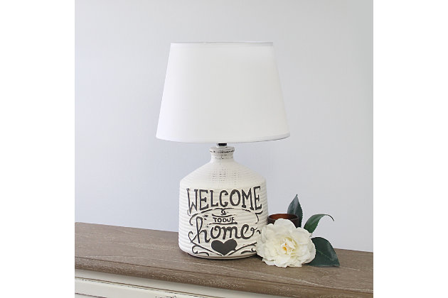 Check out this fine-looking jugular shaped table lamp spruced with a bit of a rustic appeal.  A washed out and slightly unfinished ceramic base is decorated with a black written script and paired with a white linen fabric shade.  In today's world of interior design, this lamp is the farmhouse or countryside lover's ideal lamp find.White wash ridged ceramic base with "welcome to our home" design | White linen empire fabric shade | Uses 1 x 60w type a medium base bulb (not included) | Easy on/off switch located on socket